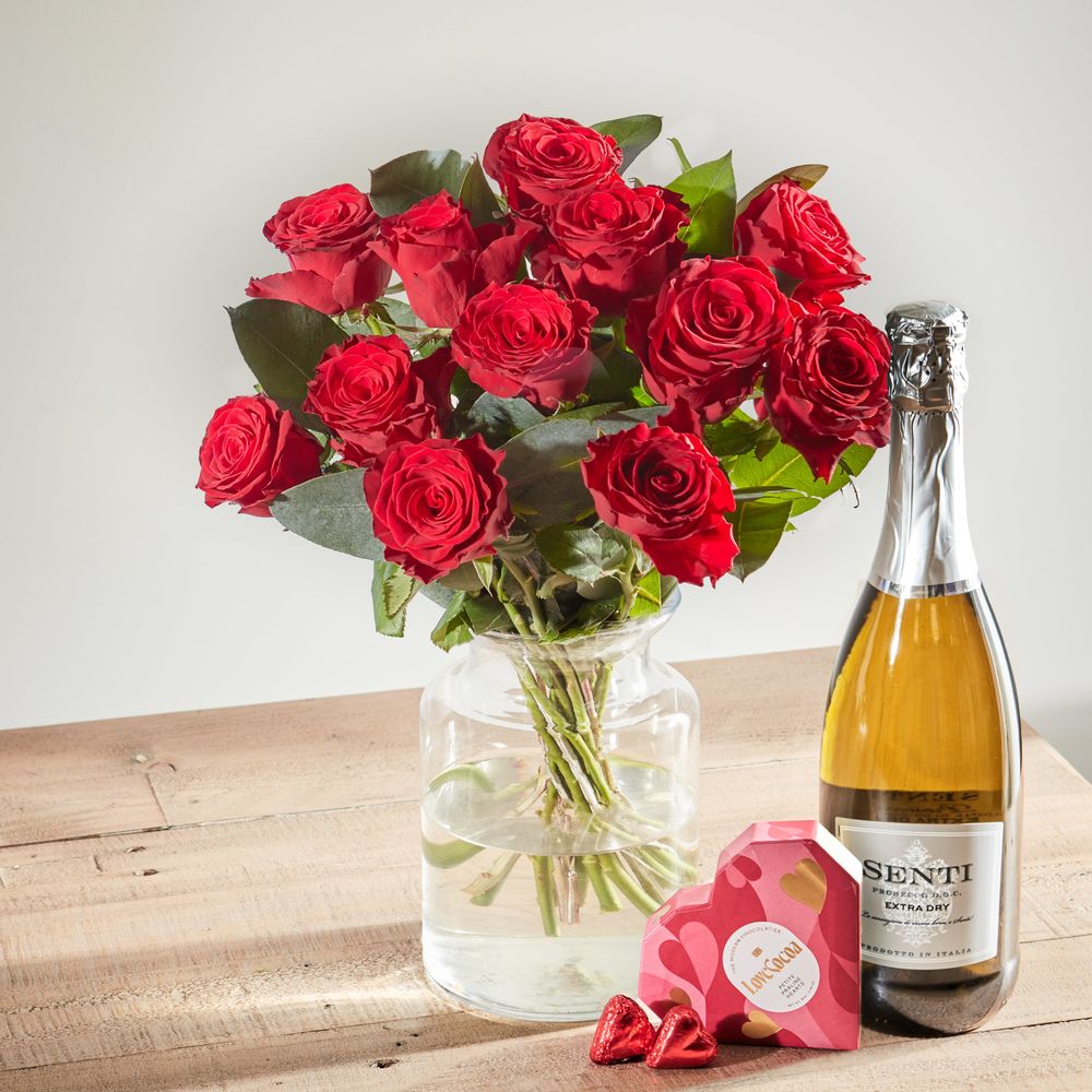 A bouquet of red roses and green eucalyptus foliage in a clear glass vase next to a deep green bottle of prosecco