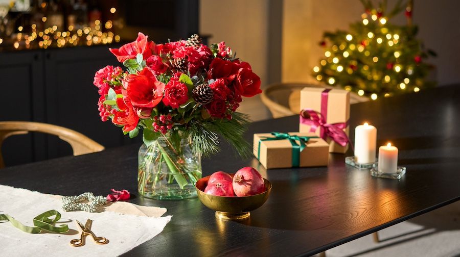 A red bouquet of amaryllis, hypericum and pine cones sits on a table with Christmas presents
