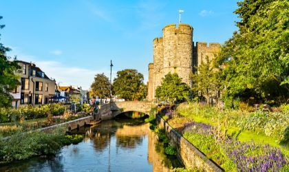 Photo of a small river on the outskirts of an old castle. There are wildflowers growing on both sides of the river, and a footbridge links the castle to the main town.