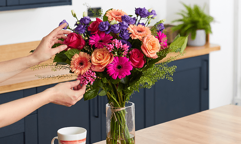 A bouquet of bright pink, coral, red and purple flowers being primped by hand on the left hand side.