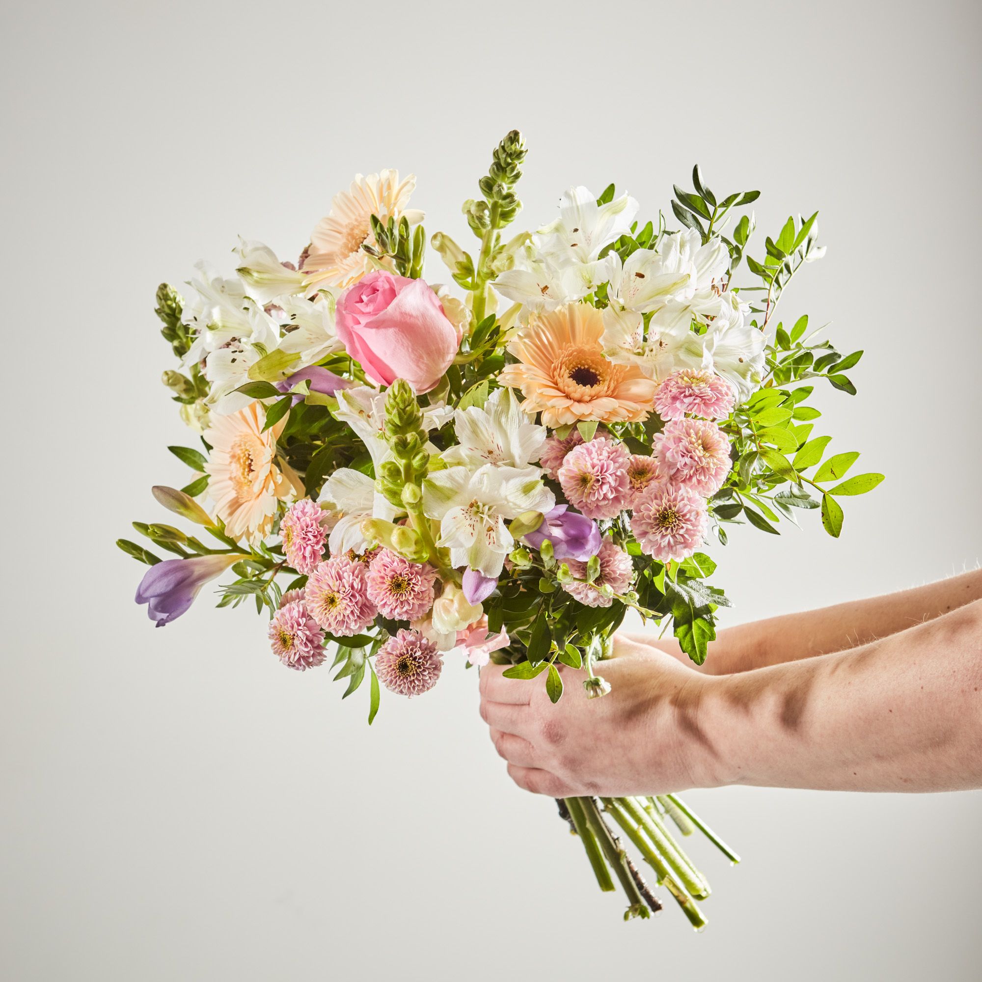 A pair of hands holds a bouquet of flowers into frame. The bouquet is a mix of pastel peach, pink and white.