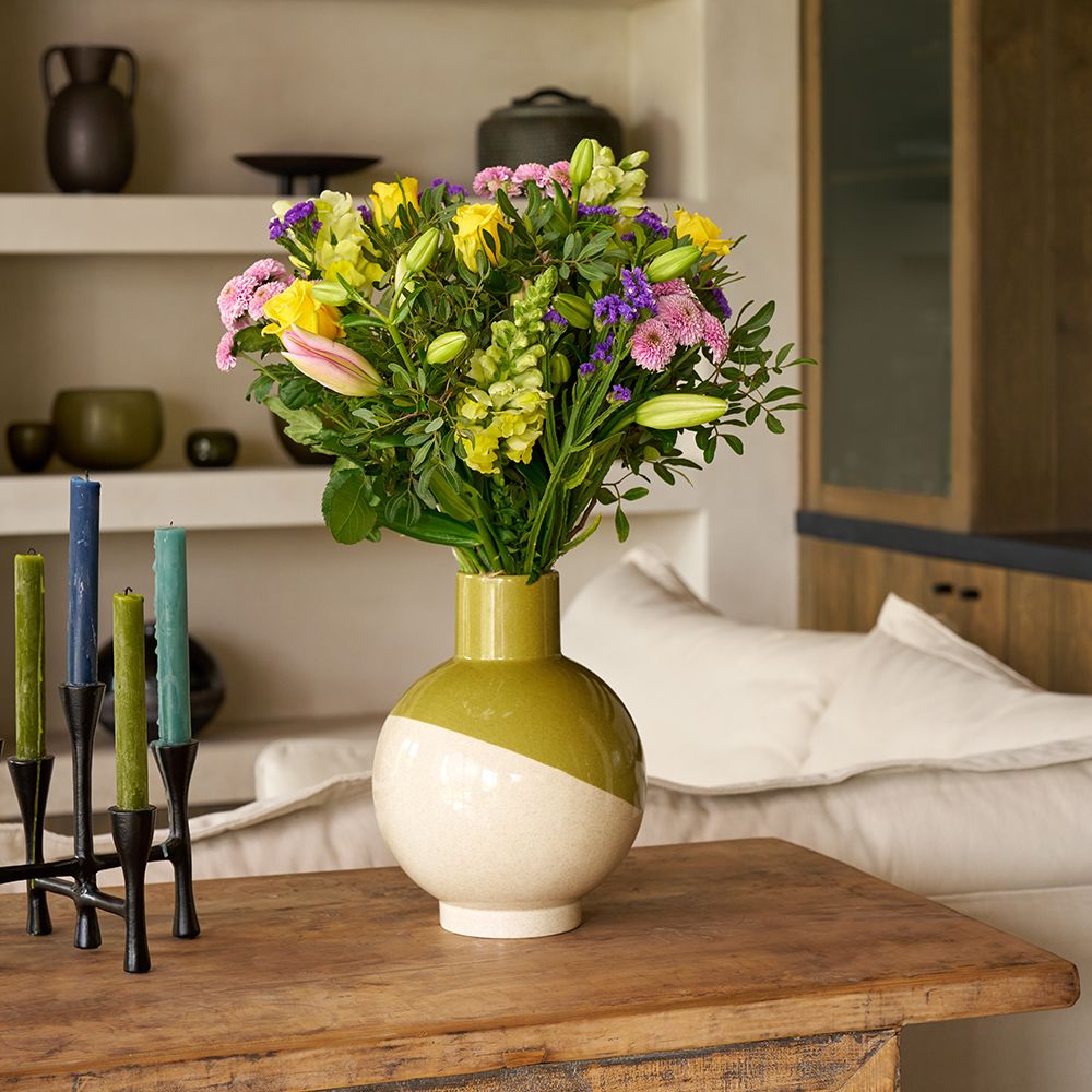 A bouquet of yellow, pink and purple flowers in a green and white vase, on a wooden sideboard