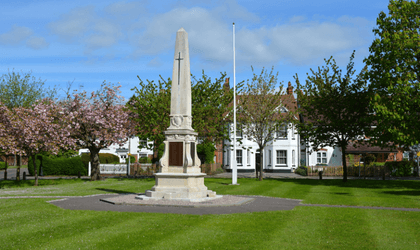 Picture of a cenotaph-style war memorial in the middle of a well manicured green and surrounded by trees in blossom.