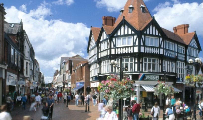 A busy high street on a sunny day. The main building in the image as a white Tudor-style house with black beams and gables that has been built with a rounded front to it fits a sharp corner.