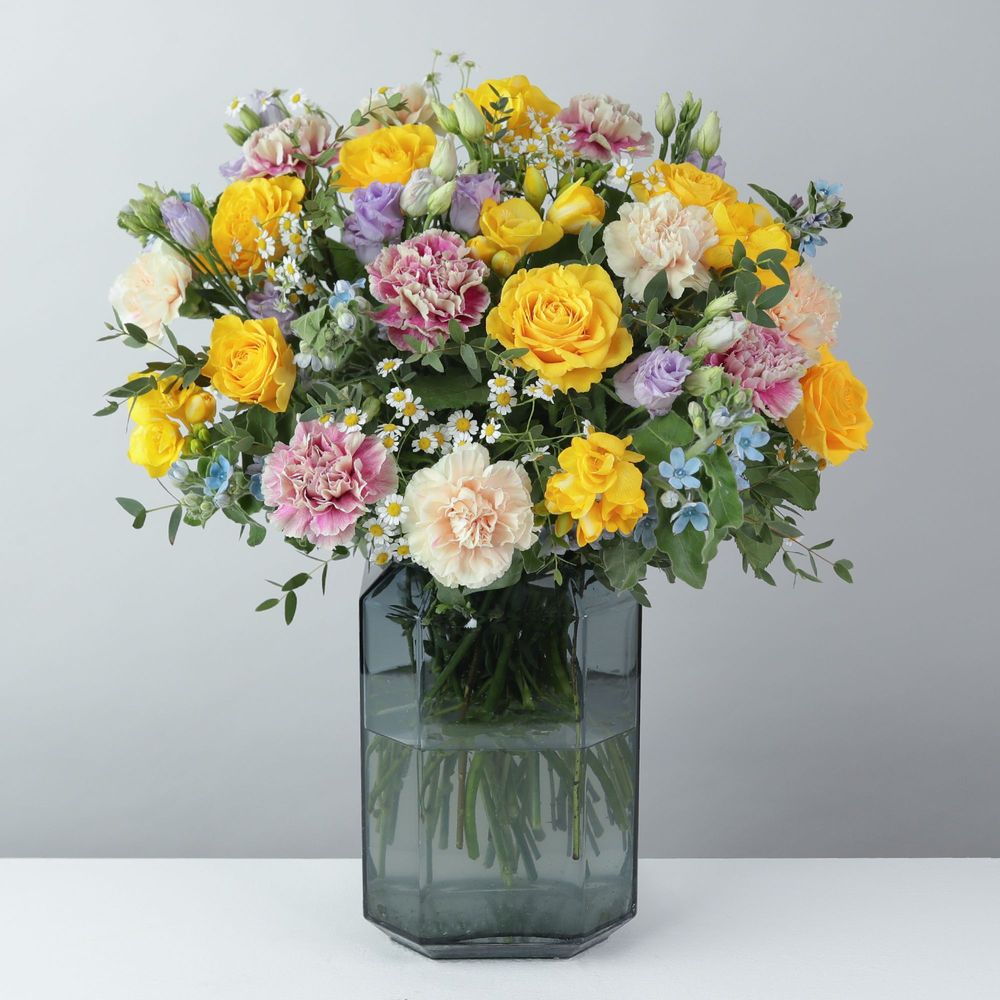 A bouquet of Yellow Roses, Blue Oxypetalum, pink Carnations, Parvi Eucalyptus, Lilac Lisianthus, Tanacetum Vegmo, cream Carnations, Yellow Freesias in a blue smoke vase that is a hexagon shape.