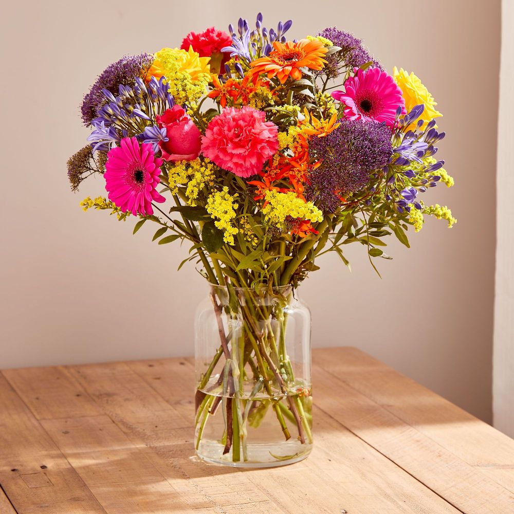 A bouquet of bright pink Gerberas, coral carnations, pink and yellow roses, lilac Agapanthus, yellow Solidago, purple Trachelium, orange Asclepias and pistache foliage. The bouquet sits in a clear round vase on a wooden country kitchen table.