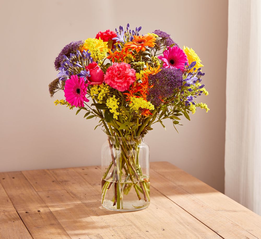 Photo of a bouquet of brightly coloured flowers. Stems include hot pink and orange gerberas, vivid pink carnations, cerise and yellow roses, orange asclepias, yellow solidago, purple trachelium, lilac agapanthus, and green pistache foliage.