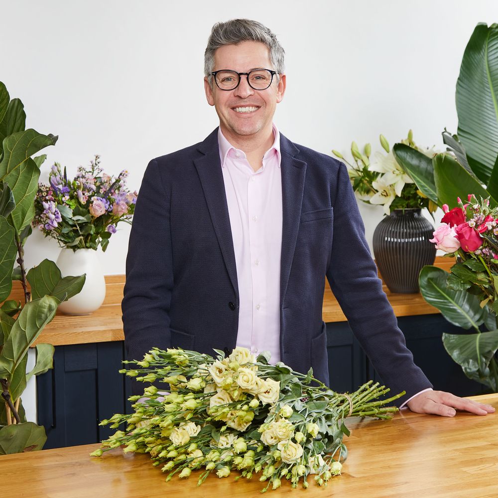 A photograph of a well-dressed man wearing a pink shirt, deep navy blazer and dark-framed glasses. The man, who is smiling, is standing at a counter with a clean and shiny wooden surface, which has a hand-tied bouquet of creamy yellow flowers on it. Surrounding him are bouquets and plants. 