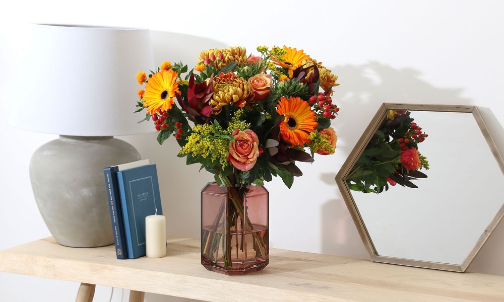 A bouquet on a shelf with a lamp, mirror, candle and two books propped up against the lamp. Flowers in the bouquet are orange ombre Gerberas, red and yellow ombre Chrysanthemums, red hypericum, yellow solidago, cherry brandy roses, deep red Leucadendron, and orange safflower.