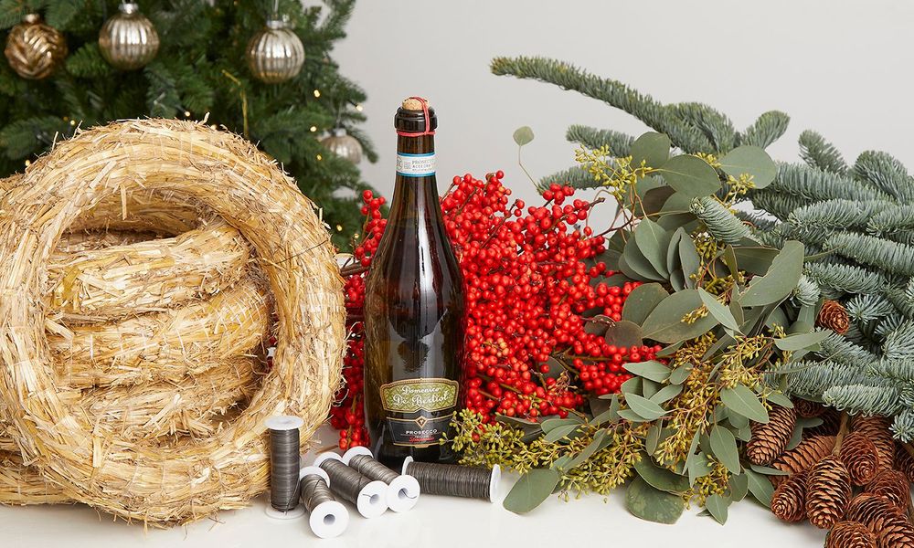 Photo of a wreath making kit featuring straw wreath bases, wire, berries, pine, eucalyptus and pine cones, with a bottle of Prosecco. 