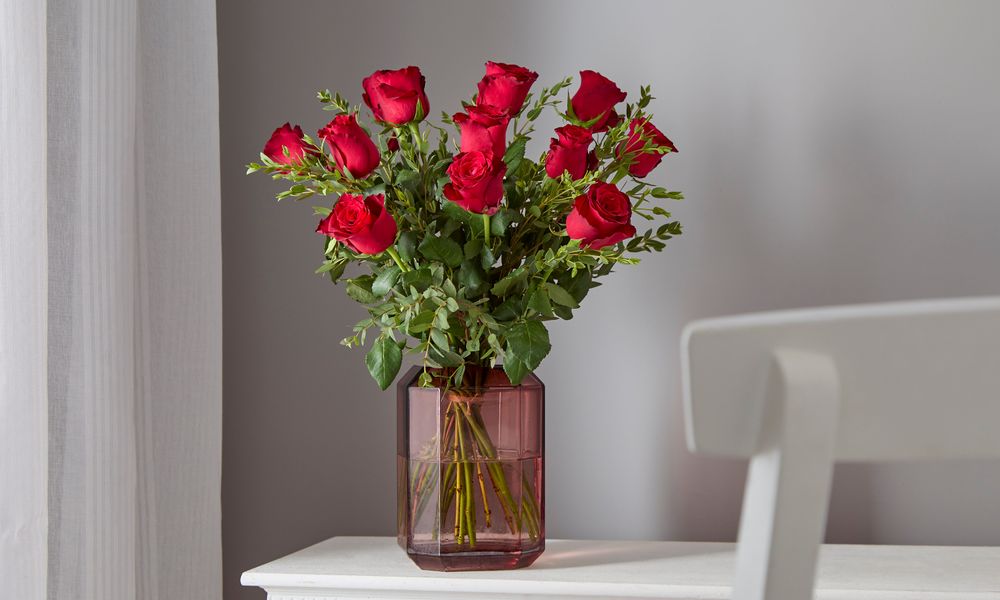 Photograph of a bouquet of 12 fairtrade red roses in a smoked pink hexagonal vase. The vase is on a white sideboard with a soft translucent curtain to its left. 