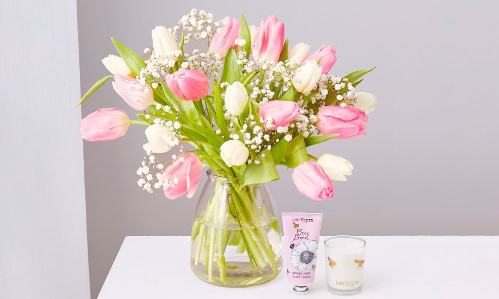 A bouquet of pink and white tulips with white Gypsophila in a clear vase. It's on a white table white a tube of 'Bee loved' hand cream and a votive candle in a glass holder with two golden bee silhouettes on it. 