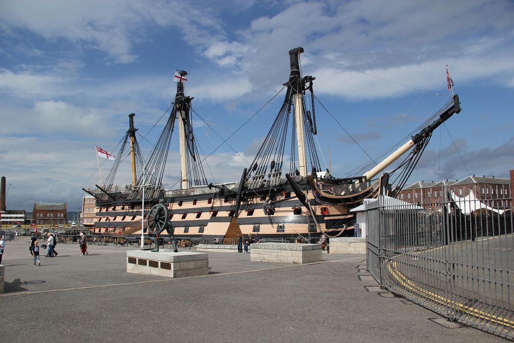 An eighteenth to nineteenth century naval vessel in Portsmouth harbour on a sunny day. The HMS Victory is perhaps the most famous warship, and is known for its role in the Battle of Trafalgar. The vessel is now a tourist attraction.