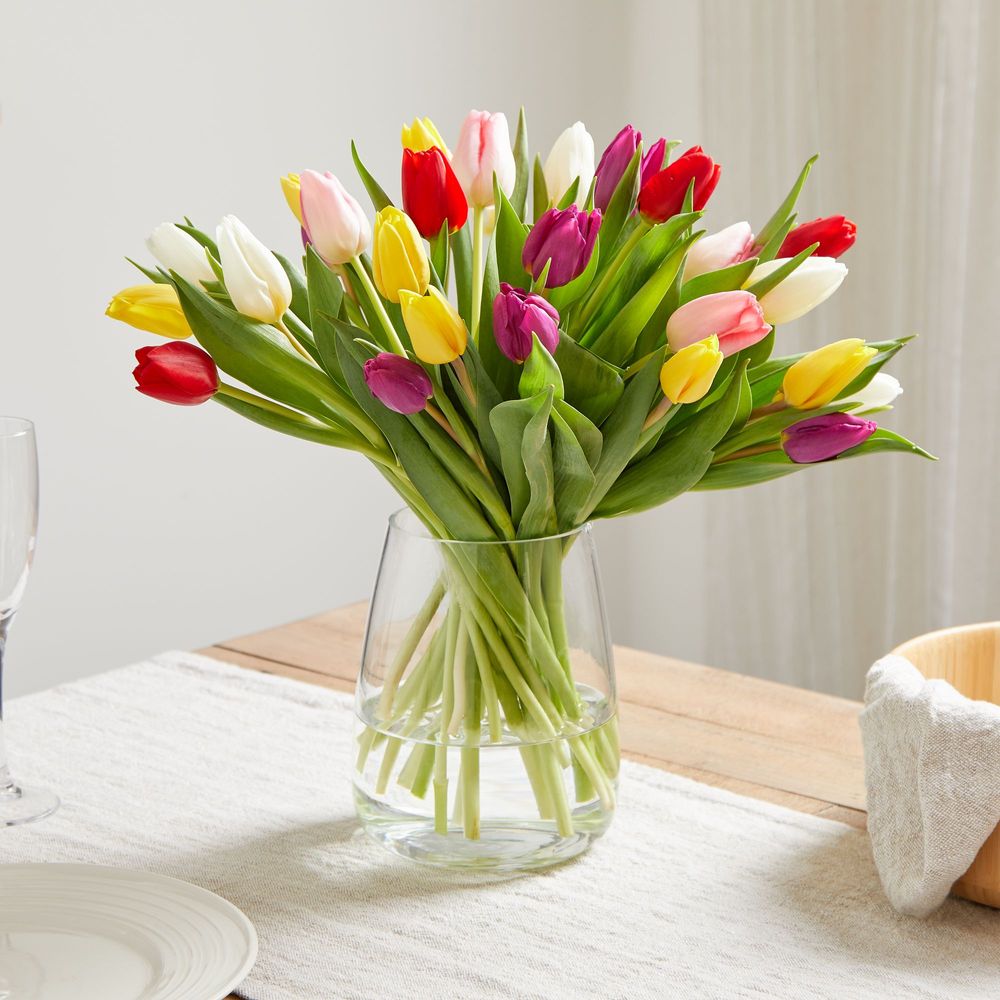 A light and airy scene of a kitchen table. On the table is a natural linen runner, simple porcelain plate, a champagne flute and a bowl. Also on the table is a simple but elegant glass vase filled with 30 tulips in an assortment of red, yellow, pink, magenta and white.