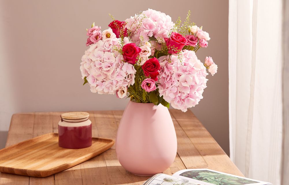 Photo of a light and airy room with a wooden kitchen table. On the table is a bamboo wooden tray with a ceramic jar, a magazine open on a page with a lush green houseplant, and a teardrop shaped pink vase. In the vase is a bouquet of delicate pale pink hydrangeas, cerise roses, bubblegum pink lisianthus, and greenbell foliage.