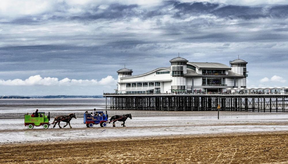 Photograph of a sandy beach, long pier and building with two carts in the style of train carriages being pulled by horses on the beach.