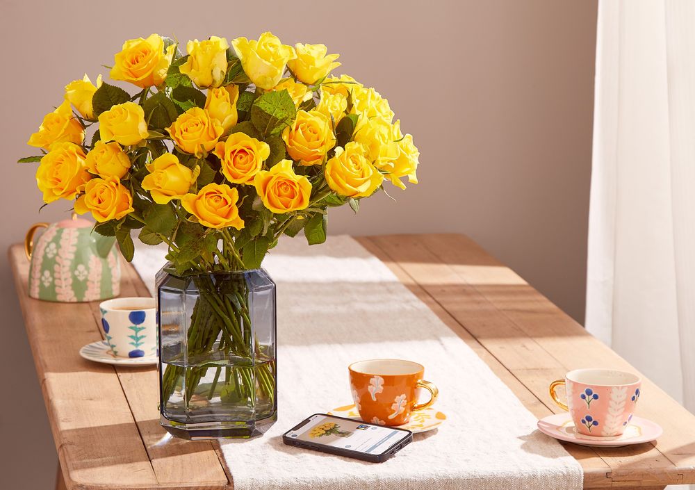 A bouquet of bright yellow roses sit in a blue smokey hexagonal vase on a wooden country kitchen table. Also on the table are three colourful cups and saucer and a teapot. Sunlight streams in through a window to the right between linen curtains.