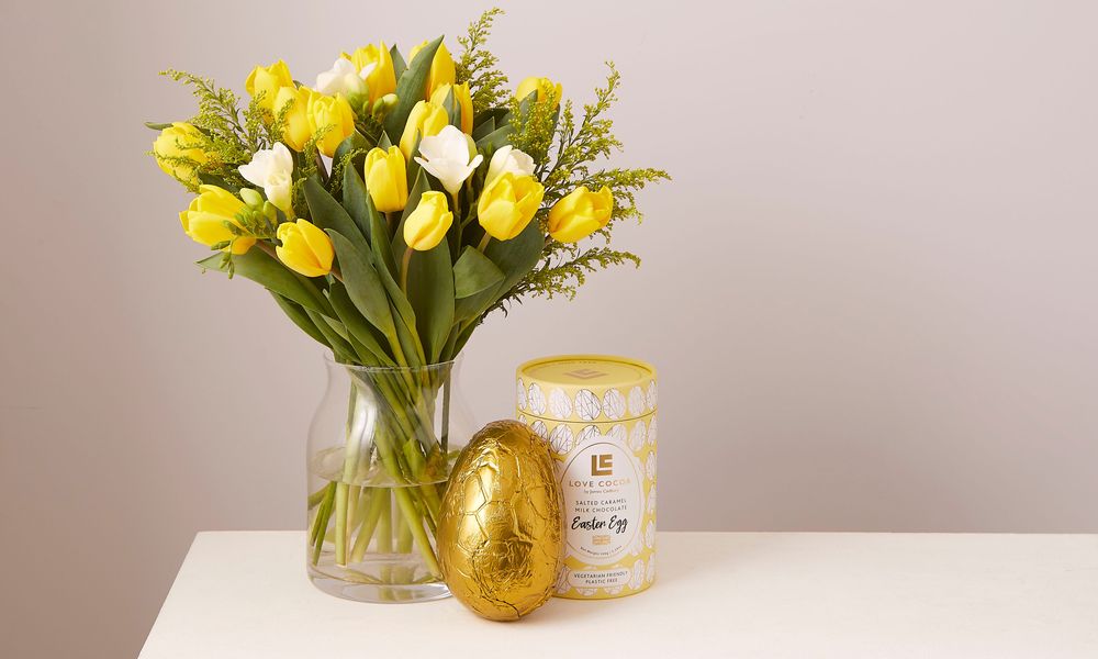A bouquet of yellow tulips, white Freesia and solidago in a clear vase on a table with a delicious looking chocolate easter egg next to it.