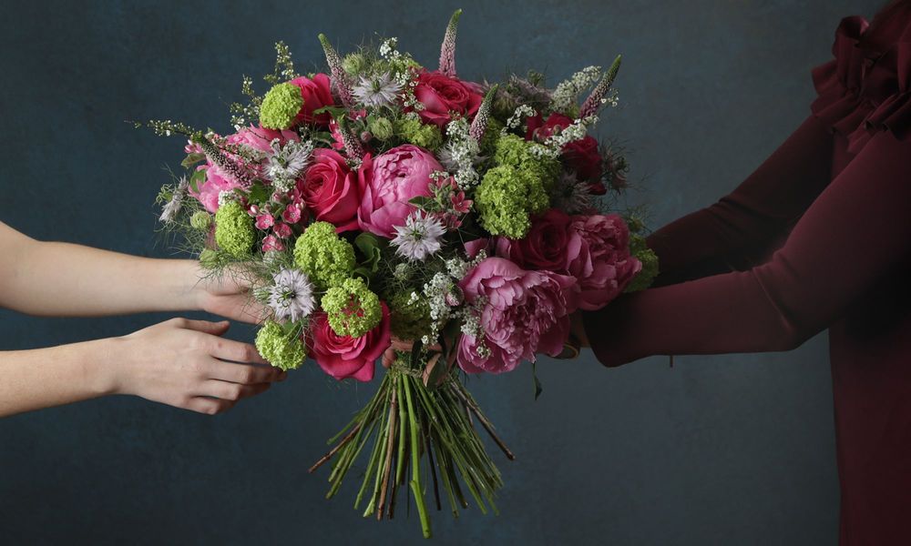 A photo of a torso on the right in a burgundy jumper handing a big, beautiful bouquet of peonies, roses, veronica, and foliage, to some hands on the left.