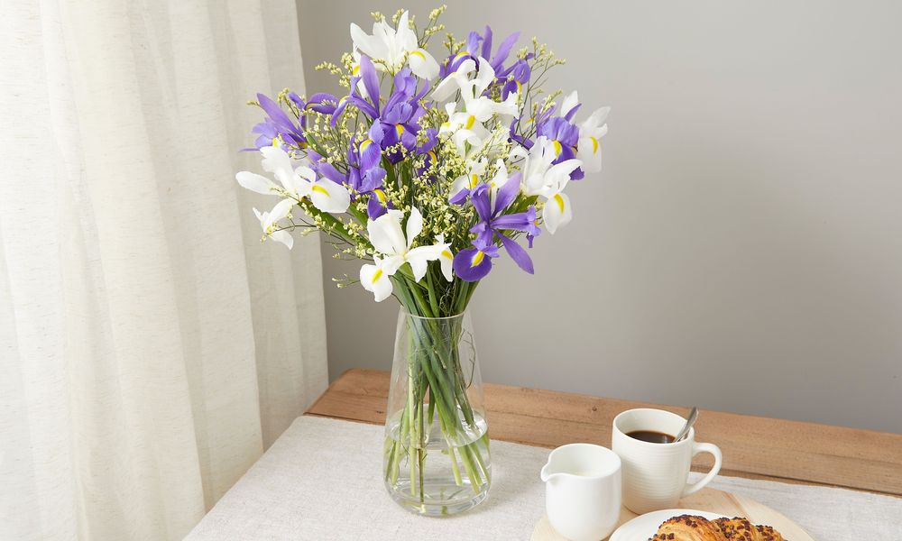 A bouquet of violet and white irises with yellow limonium sits on a wooden dining table. There's a natural linen table runner across the middle of it. A very tasty looking croissant sits on a plate next to a cup of strong coffee and milk in a jug.
