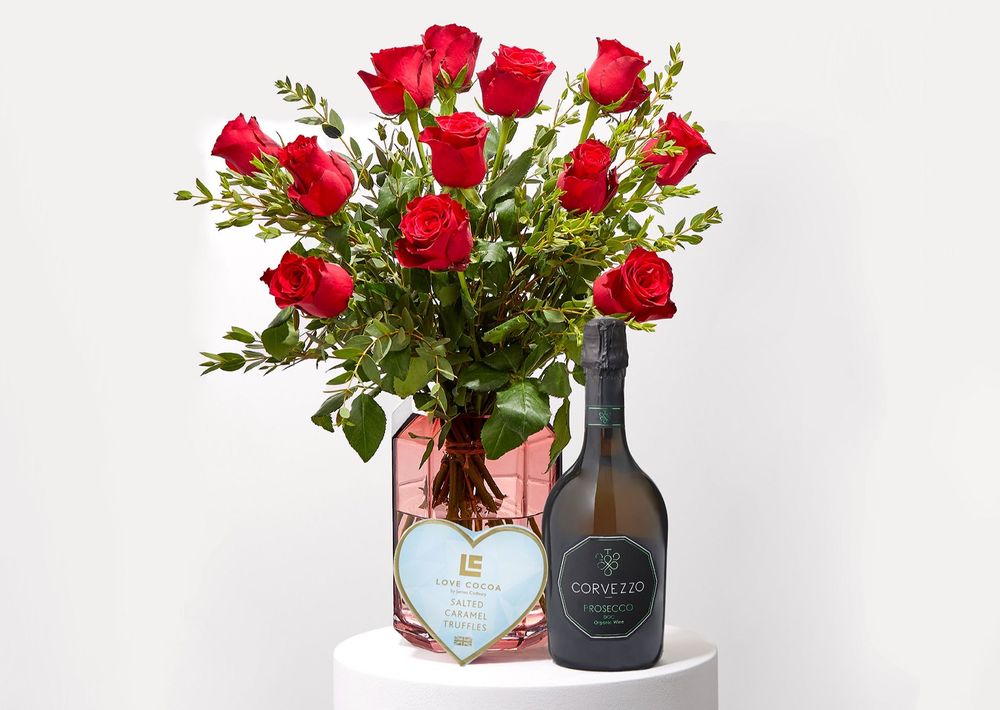 Red roses in a pink vase with a bottle of Prosecco and heart-shaped box of chocolates